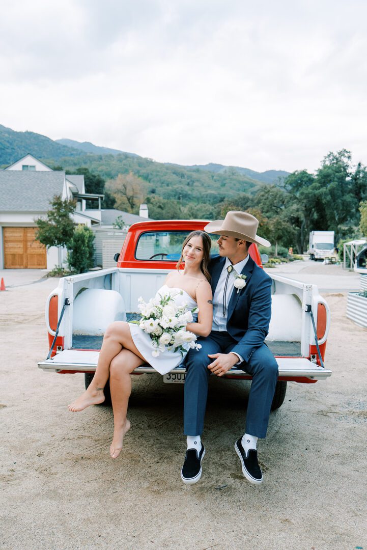 Newlywed couple sitting on the back of a vintage car, sharing a joyful moment.