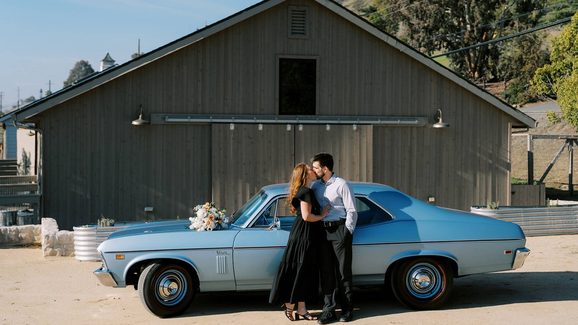 Couple embraces beside a vintage blue car in front of a rustic barn.