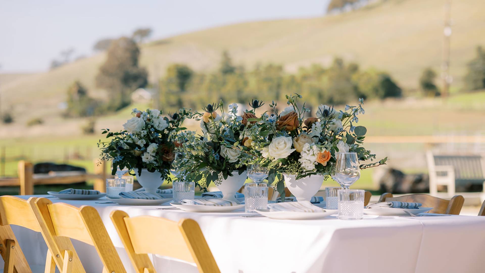 Elegant floral centerpiece on an outdoor dining table with a scenic backdrop.