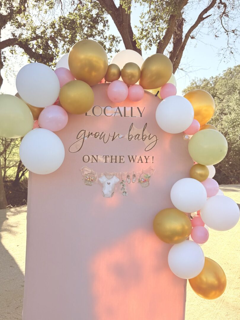 A pink and gold balloon arch with a baby shower sign.
