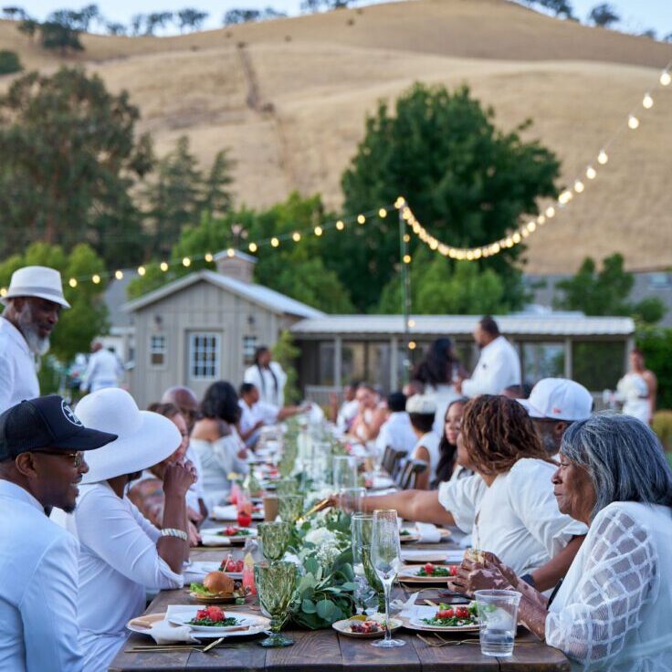 Outdoor gathering with people dining under string lights in a rural setting.